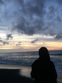 a person standing on the beach looking out at the ocean with clouds in the sky