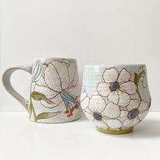 two mugs with flowers painted on them sitting next to each other in front of a white wall