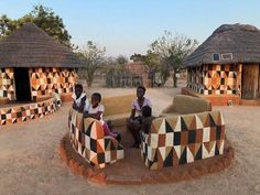 three people sitting on top of a couch in the middle of a dirt field next to huts