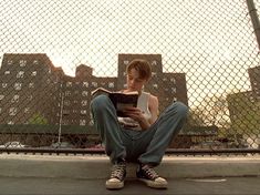 a young man sitting on the ground reading a book in front of a chain link fence