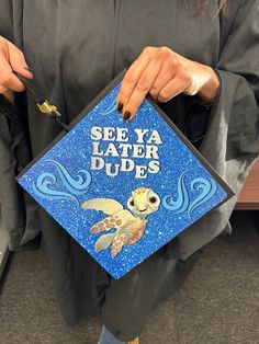 a person holding a blue graduation cap with an image of a turtle on it that says see ya later dudes