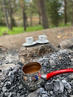 a red handled cup sitting on top of a pile of rocks next to a metal mug