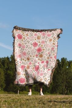 a person standing in the grass holding up a blanket with pink flowers on it and trees in the background