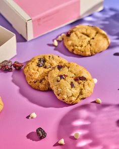 three cookies with cranberries and white chocolate chips on a purple surface next to a pink box