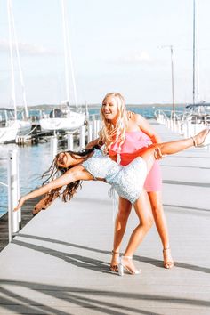 two women are dancing on a dock near the water with boats in the background and one woman is wearing a pink dress