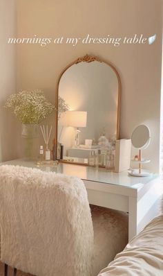 a white desk with a mirror, stool and flowers on it in front of a window