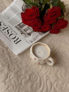 a coffee cup sitting on top of a bed next to a bouquet of red roses