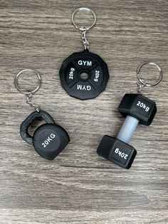 three black dumbbells and two key chains on a wooden table with the words gym written on them