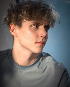 a young man with curly hair looks into the distance while wearing a gray shirt and looking off to the side