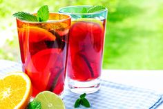 two glasses filled with red and green drinks next to sliced oranges on a table