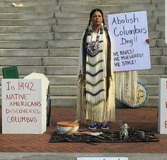 two native americans holding signs in front of some steps with other signs on them that read, abollish columbus day