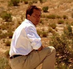 a man squatting in the desert with his hands on his hips and looking off into the distance