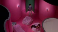 two people standing in a room with pink walls and beds on the floor, one person is taking a photo