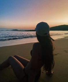 a woman sitting on top of a sandy beach next to the ocean at sun set