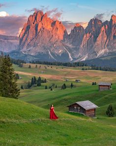 a woman in a red dress is standing on a grassy hill with mountains in the background