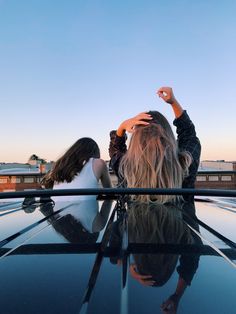 two women sitting on top of a car looking out at the sky with their arms in the air