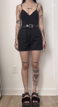 a woman with tattoos standing in front of a white wall wearing black shorts and sandals