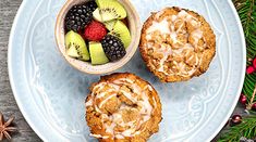 two muffins on a blue plate with fruit in the bowl and pine cones