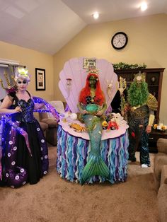 two women dressed up as mermaids sitting at a table in a living room with a clock on the wall