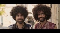 two men with curly hair and beards standing next to each other on the street