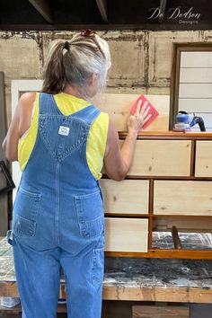 a woman in overalls standing next to a dresser and looking at something on the floor