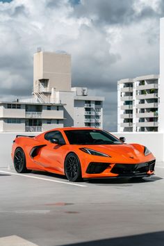 an orange sports car parked in a parking lot next to tall buildings and cloudy skies