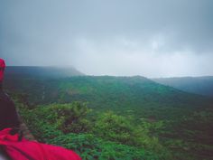 a person sitting on top of a lush green hillside covered in fog and rain while looking at the mountains