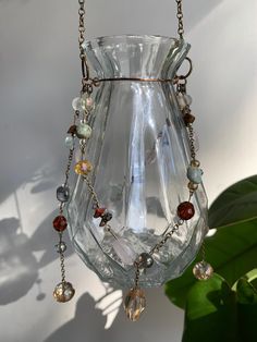 a glass vase with beads hanging from it's side and a plant in the background