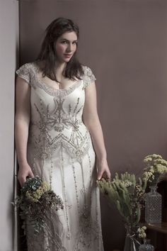 a woman in a white dress standing next to a vase with flowers and greenery