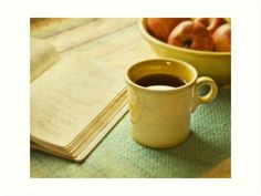 a cup of coffee sitting on top of a table next to an open book and bowl of apples