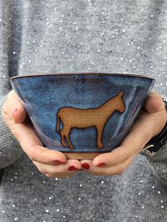 a person holding a bowl with a horse on it in their left hand and the other hand over her shoulder