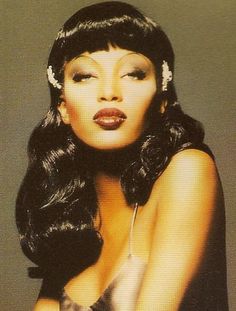 a woman with long black hair wearing a silver dress and white headband, posing in front of a gray background