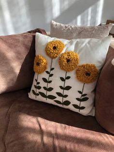 a pillow with three yellow flowers on it sitting on a brown couch in front of a window