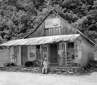 black and white photograph of a man sitting in front of a small shack with a dog