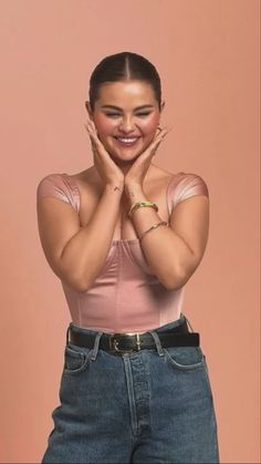 a woman with her hands on her face posing for the camera while wearing jeans and a pink top