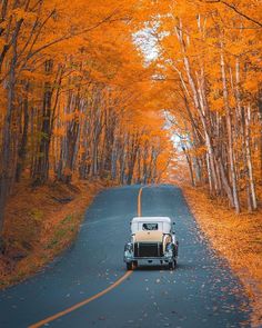 an old car driving down the road in front of trees with orange leaves on them