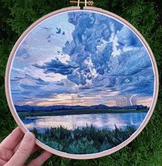 a hand holding up a cross stitched picture with clouds and water in the background