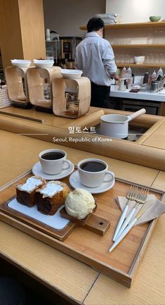 two cups of coffee and some cake on a wooden tray in a kitchen with a person behind the counter