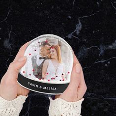 a hand holding a snow globe with a couple in it and hearts on the inside