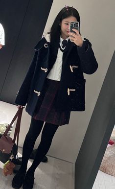 a woman taking a selfie in front of a mirror wearing a black coat and plaid skirt