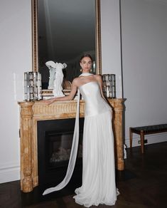 a woman standing in front of a fireplace wearing a white dress and holding a long veil