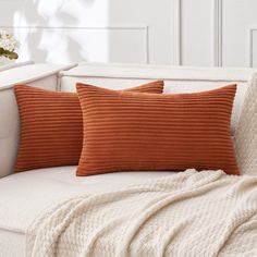 two orange pillows sitting on top of a white couch next to a blanket and flowers