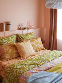 a bed with colorful sheets and pillows next to a window in a pink walled room