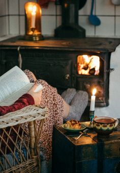 a woman sitting in a chair reading a book next to an open fire place with food on the table