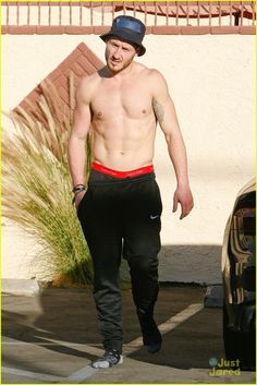 a shirtless man walking down the street wearing black pants and a hat with tattoos on his arm