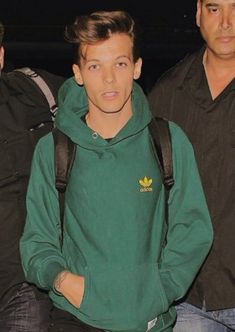 the man is wearing a green hoodie and black pants with his hands in his pockets