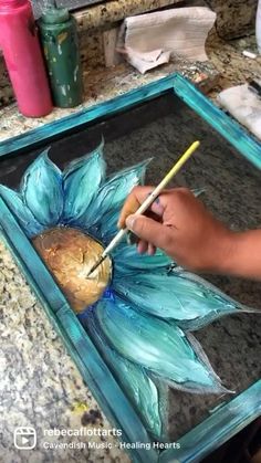 a person is painting a flower with blue paint on the glass tray and holding a pencil in their left hand