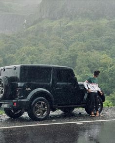 two people standing next to a black jeep on the side of a road in the rain