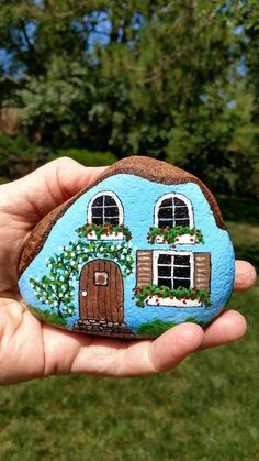 a hand holding a painted rock with a house on it in front of some trees