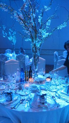 a table set up for a formal dinner with blue lighting and flowers in the center